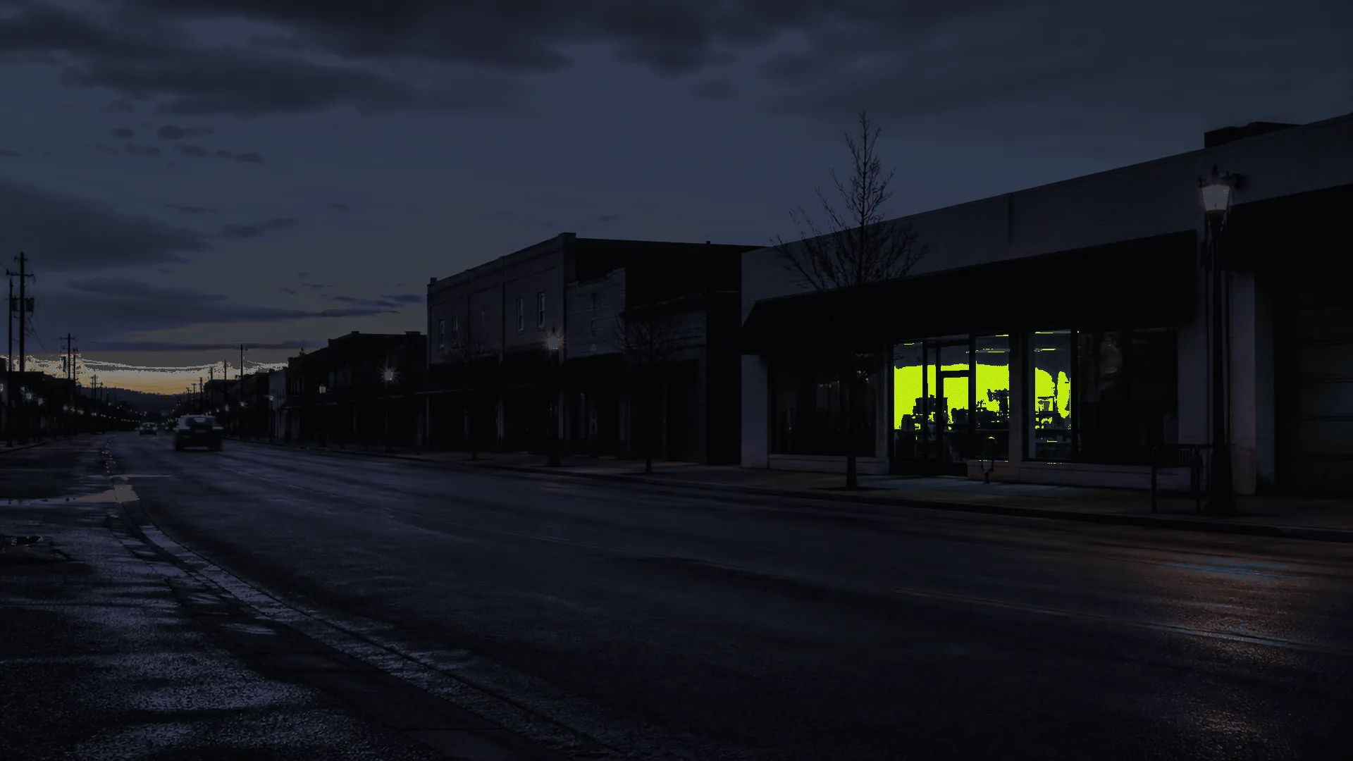 Dusk on a quiet service-business main street emptying out — one storefront still lit by a restrained accent of practical light.